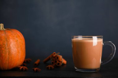 Glass mug of coffee with milk and foam, cinnamon spice sticks, pumpkin on black table against dark blue wall background. Autumn drink cup, fall cocoa, spicy latte, thanksgiving, coffee shop. Cocoa