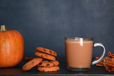 Süt ve köpüklü bir bardak kahve, tarçın, kurabiye, koyu mavi duvar arka planında siyah masada turuncu balkabağı. Sonbahar içkisi konsepti. Sonbahar, baharatlı latte, şükran günü, kafe menüsü, kapat.