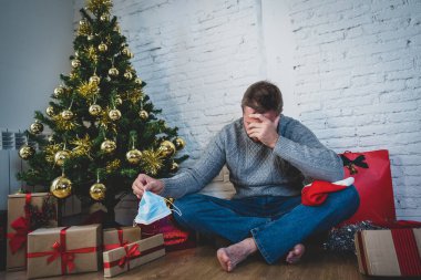 Maskeli üzgün bir adam Noel 'de yalnız başına evde yalnız başına yas tutuyor ve ailesini ve arkadaşlarını özlüyor. COVID-19 Kış tatili ve akıl sağlığı süresince karantina ve tecrit.