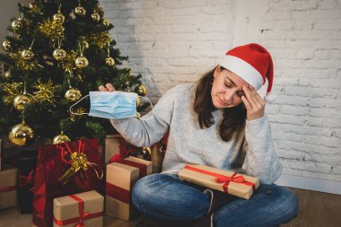 Maskeli üzgün kadın Noel 'de yalnız başına evde yalnız başına yas tutuyor ve ailesini ve arkadaşlarını özlüyor. COVID-19 Kış tatili ve akıl sağlığı süresince karantina ve tecrit.