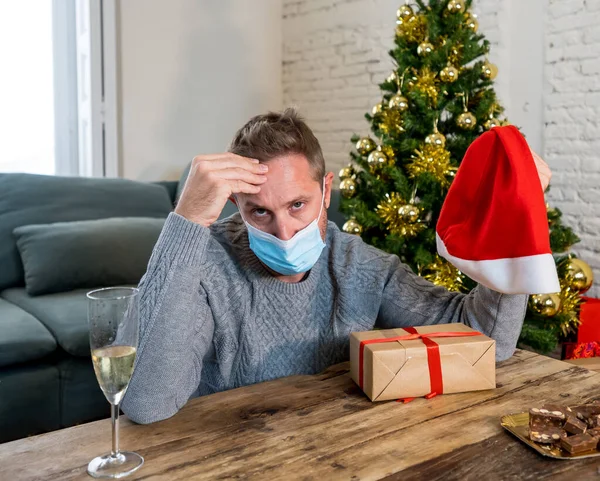 Sad man with mask home alone in self isolation at christmas feeling depressed mourning and missing family and friends. COVID-19 Lockdown and quarantine during winter holidays and mental health.