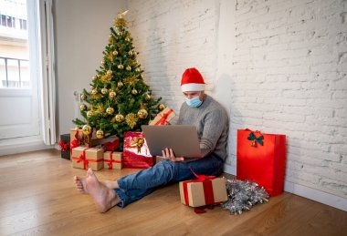 Yüzü maskeli mutlu adam evde yalnız başına sanal Noel videosunu kutluyor ailesini ve arkadaşını arıyor. Coronavirüs nedeniyle sanal tatil çevrimiçi Yeni normal ve sosyal mesafeler.