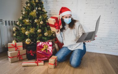 Yüzü maskeli mutlu bir kadın evde yalnız başına Noel 'i kutluyor. İnternetten ailesini ve arkadaşlarını arıyor. İkinci koronavirüs salgını nedeniyle sanal tatil ve kutlamalar.
