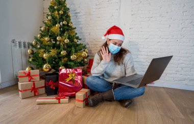 Yüzü maskeli mutlu bir kadın evde yalnız başına Noel 'i kutluyor. İnternetten ailesini ve arkadaşlarını arıyor. İkinci koronavirüs salgını nedeniyle sanal tatil ve kutlamalar.