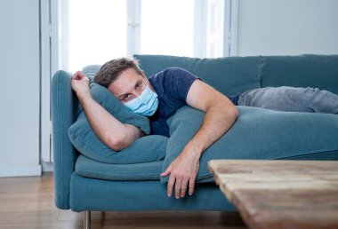 Oturma odasındaki kanepede koruyucu maskeli üzgün bir adam. Koronavirüs kilitlenmesi ve sosyal mesafeler arasında bunalıma girmiş yorgun ve endişeli hissediyor. Ruh sağlığı ve tecrit kavramı.