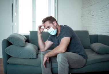 Oturma odasındaki kanepede koruyucu maskeli üzgün bir adam. Koronavirüs kilitlenmesi ve sosyal mesafeler arasında bunalıma girmiş yorgun ve endişeli hissediyor. Ruh sağlığı ve tecrit kavramı.