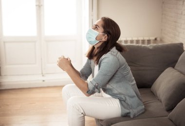 Oturma odasındaki kanepede koruyucu maskeli üzgün Latin kadın. Koronavirüs hapsi ve sosyal mesafeler yüzünden bunalımda ve yorgun hissediyor. Ruh sağlığı ve tecrit kavramı.