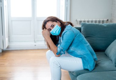 Oturma odasındaki kanepede koruyucu maskeli üzgün Latin kadın. Koronavirüs hapsi ve sosyal mesafeler yüzünden bunalımda ve yorgun hissediyor. Ruh sağlığı ve tecrit kavramı.