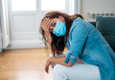 Oturma odasındaki kanepede koruyucu maskeli üzgün Latin kadın. Koronavirüs hapsi ve sosyal mesafeler yüzünden bunalımda ve yorgun hissediyor. Ruh sağlığı ve tecrit kavramı.