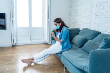 Oturma odasındaki kanepede koruyucu maskeli üzgün Latin kadın. Koronavirüs hapsi ve sosyal mesafeler yüzünden bunalımda ve yorgun hissediyor. Ruh sağlığı ve tecrit kavramı.