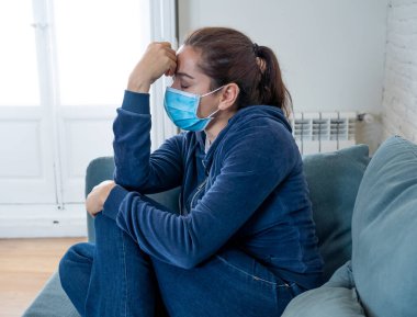 Oturma odasındaki kanepede koruyucu maskeli üzgün Latin kadın. Koronavirüs hapsi ve sosyal mesafeler yüzünden bunalımda ve yorgun hissediyor. Ruh sağlığı ve tecrit kavramı.