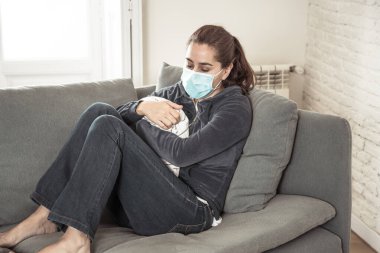 Oturma odasındaki kanepede koruyucu maskeli üzgün Latin kadın. Koronavirüs hapsi ve sosyal mesafeler yüzünden bunalımda ve yorgun hissediyor. Ruh sağlığı ve tecrit kavramı.