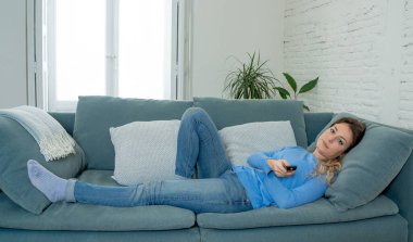Koronavirüs salgını nedeniyle zorunlu tecrit sırasında evde çaresizce televizyon izleyen sıkıcı kadın. Kanepede oturan üzgün genç kadın uzaktan kumandayla TV 'den ve sabit hayattan sıkılıyor..