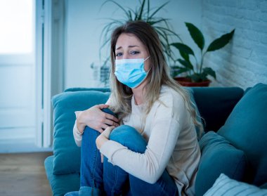 Oturma odasındaki kanepede koruyucu maskeli üzgün, yorgun ve endişeli bir kadın depresyon geçiriyor. Koronavirüs salgınından ve izolasyon konseptinden etkilenen akıl sağlığı.