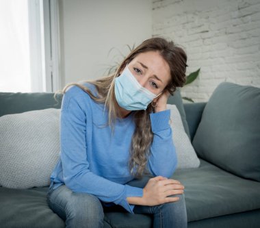 Oturma odasındaki kanepede koruyucu maskeli üzgün, yorgun ve endişeli bir kadın depresyon geçiriyor. Koronavirüs salgınından ve izolasyon konseptinden etkilenen akıl sağlığı.