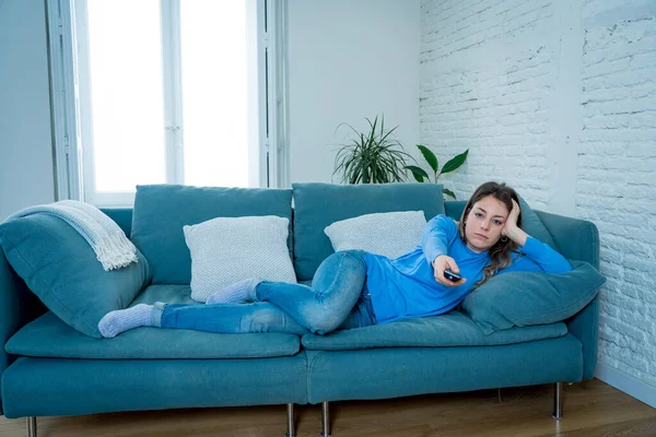 Bored woman watching TV helpless in self Isolation at home during mandatory lockdown due to coronavirus outbreak. Young upset woman on sofa using control remote zapping bored of TV and sedentary life.