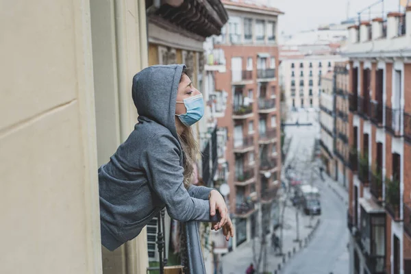 Beautiful depressed lonely woman with face mask in quarantine staring out on balcony feeling sad, lonely, pain and grief during coronavirus outbreak. Depression and mental health due to COVID-19.