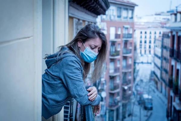 Beautiful depressed lonely woman with face mask in quarantine staring out on balcony feeling sad, lonely, pain and grief during coronavirus outbreak. Depression and mental health due to COVID-19.