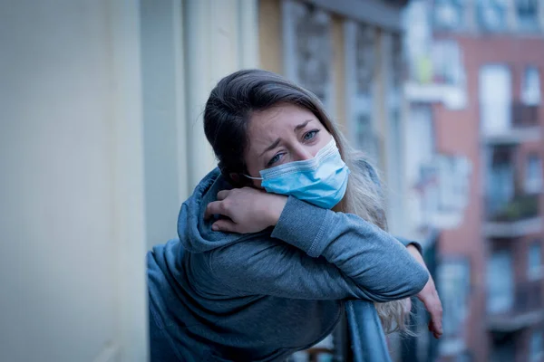 Beautiful depressed lonely woman with face mask in quarantine staring out on balcony feeling sad, lonely, pain and grief during coronavirus outbreak. Depression and mental health due to COVID-19.