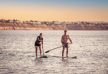 SUP kürekli sörf tahtasında, gün batımında sakin denizde güzel kumsalda eğlenen olgun beyaz bir çift. Aktif modern yaşlı insanlar, açık hava aktiviteleri, birlikte yaşlanma ve sağlıklı yaşam tarzı..