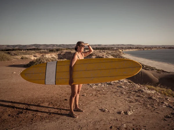 Attractive Australian mature surfer with cool vintage surfboard at sunset. Fit Woman in her 50s happy to be Back to surf . Outdoors sports adventure, active older people and retirement lifestyle.