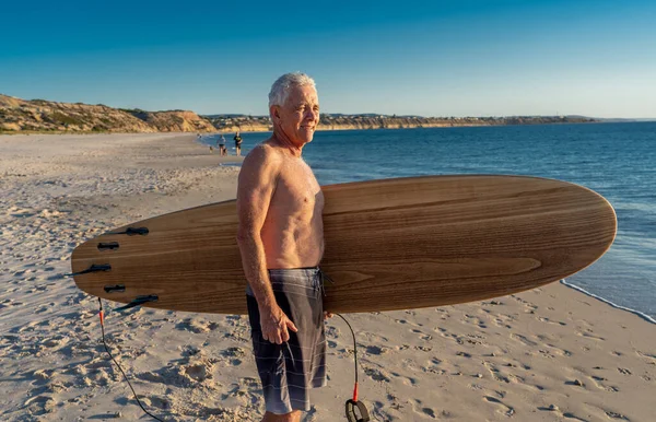 Attractive Australian mature man surfer with cool vintage surfboard on beach at sunset. Senior adult happy to be Back to surf . Outdoors sports adventure, active older people and retirement lifestyle.