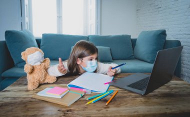E-öğrenme. Sıkıcı ve depresif kız, oyuncak ayı ve yüz maskesi takmış evde online derslerde ders çalışıyor çünkü yeni COVID-19 hava durumu ya da hava tahminleri nedeniyle okul kapalı..
