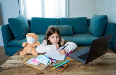 E-öğrenme. Sıkıcı ve depresif kız, oyuncak ayı ve yüz maskesi takmış evde online derslerde ders çalışıyor çünkü yeni COVID-19 hava durumu ya da hava tahminleri nedeniyle okul kapalı..
