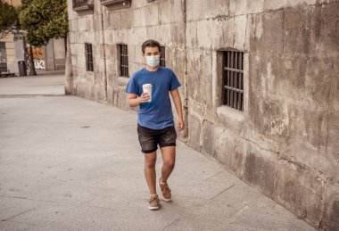 COVID-19 salgını. Şehir sokaklarında koruyucu cerrahi maske takmış ve kahve içen genç bir adam. Coronavirus, Yeni Normal ve zorunlu yüz maskesi kullanımı
