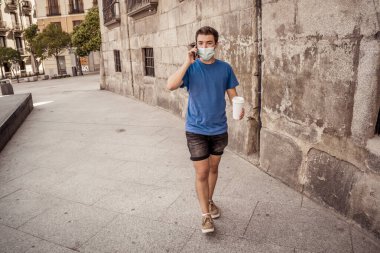 COVID-19 salgını. Şehir sokaklarında koruyucu cerrahi maske takmış ve kahve içen genç bir adam. Coronavirus, Yeni Normal ve zorunlu yüz maskesi kullanımı