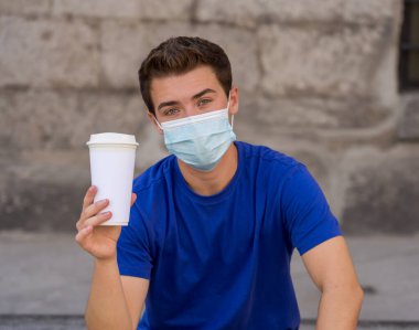 COVID-19 salgını. Şehir sokaklarında koruyucu cerrahi maske takmış ve kahve içen genç bir adam. Coronavirus, Yeni Normal ve zorunlu yüz maskesi kullanımı