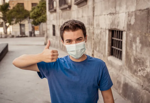 Happy young man with surgical face mask outdoors showing thumbs up. Man with protective mask walking in city street after coronavirus outbreak lockdown. Positive image New Normal life and COVID-19.