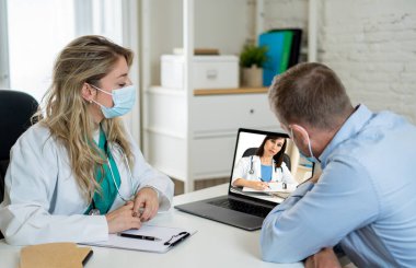 Klinikte yüz maskesi takan kadın doktor ve erkek hasta sağlık kliniğinde ya da hastanede online doktor uzmanıyla konuşuyor. Çevrimiçi Tıp, Teknoloji ve Sanal Tıbbi Danışmanlık.