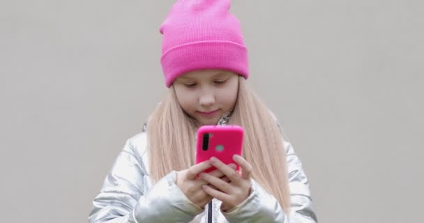 Una colegiala con un sombrero rosa sostiene un teléfono móvil en sus manos y escribe el mensaje. — Vídeos de Stock