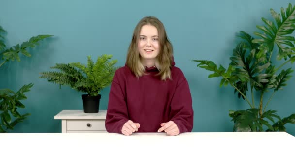 Een schattig meisje zit aan een tafel tegen een achtergrond van groene planten. — Stockvideo