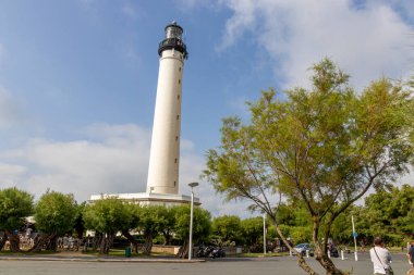 Biarritz, Fransa. Hafif bulutlu bir yaz gökyüzünün altında çevre bahçe bitkisinin üzerinde yükselen beyaz deniz fenerinin tam görüntüsü