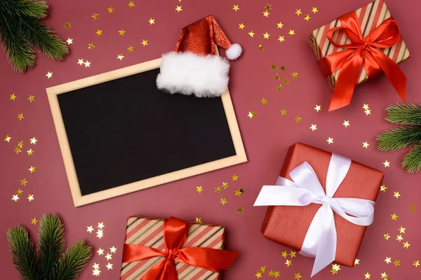 Açık ahşap çerçeve, yazı için siyah yer, Noel Baba şapkası. Yılbaşı dalları. Kırmızı arka planda Xmas hediyeleri ve altın yıldızlar. Flatlay. Uzayı kopyala Hiç kimse. Noel kartı..