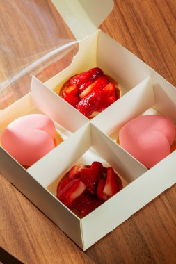 Kalp şeklinde kremalı pasta seti ve içinde çilek olan kurabiye sepetleri. Kafedeki bir masada üzerinde kekler olan karton kutu, seçici bir odak.