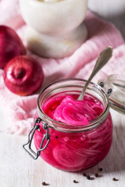 Cam kavanozdaki parlak pembe turşulu kırmızı soğanlar, taze soğan ve baharatlarla kırsal bir arka planda servis ediliyor. Sağlıklı beslenme, vegan yemek tarifleri, temiz yemek blogları, konserve, muhafaza ve organik yaşam temaları için ev yapımı mayalanmış sebze konsepti.