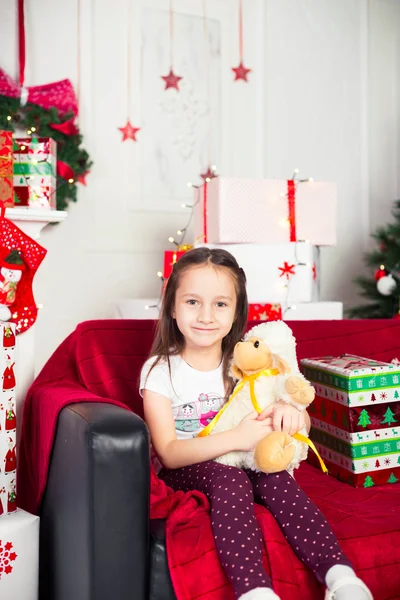 Menina sentada no treinador segurando brinquedo de pelúcia, presente de Natal, bandeja no fundo — Fotografia de Stock
