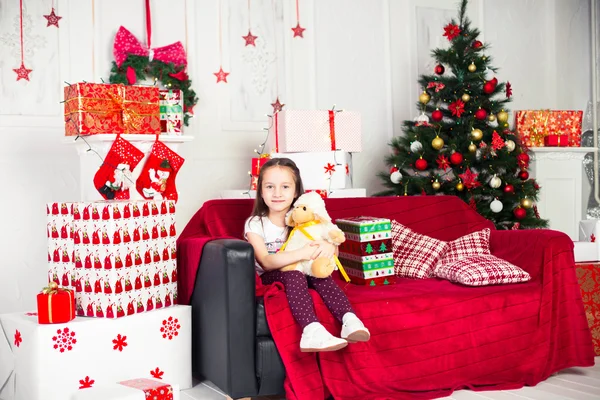 Menina sentada no treinador segurando brinquedo de pelúcia, presente de Natal, bandeja no fundo — Fotografia de Stock