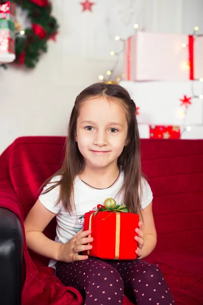 Little girl sitting on couch holding red box, gift, Christmas tree in the background — Stock Photo, Image