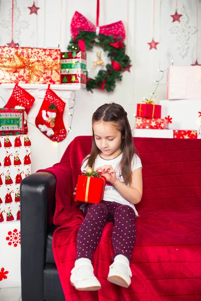 Little girl sitting on couch holding red box, gift, Christmas tree in the background — Stock Photo, Image