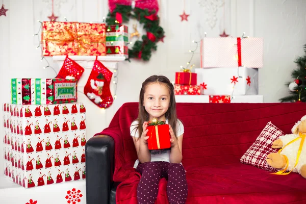 Menina sentada no sofá segurando caixa vermelha, presente, árvore de Natal no fundo — Fotografia de Stock