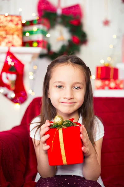 Little girl sitting on couch holding red box, gift, Christmas tree in the background — Stock Photo, Image