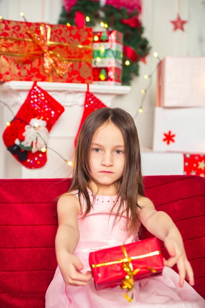 Little girl sitting in ping dress holding red box, gift, Christmas tree on background — Stock Photo, Image