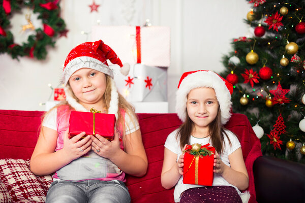 Two girls Christmas costumes sitting on the sofa with gifts in hand, tree background