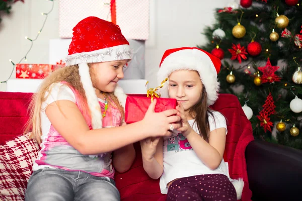 Two girls in Christmas costumes sitting on the couch give each other gifts, tree background — Stock Photo, Image