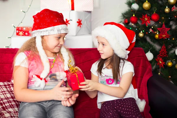 Duas meninas em trajes de Natal sentadas no sofá dão presentes um ao outro, fundo da árvore — Fotografia de Stock