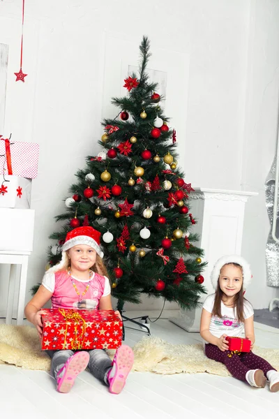 Duas meninas em trajes de Natal sentadas sob a árvore com presentes suas mãos — Fotografia de Stock